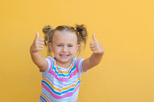 portrait happy surprise little kid girl in colored t-shirt showing point fingers thumbs up hands smiling recommend isolated over yellow color background