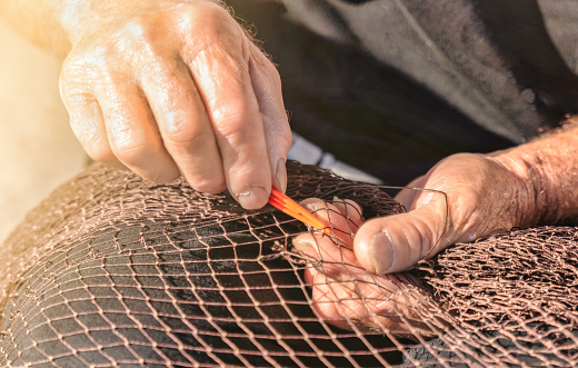 Senior fisherman repairs a fishnet by hand with a needle - Traditional fishing industry concept.
