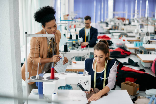 istock Young seamstress sewing clothes while production line manager is overseeing her. 1409861285