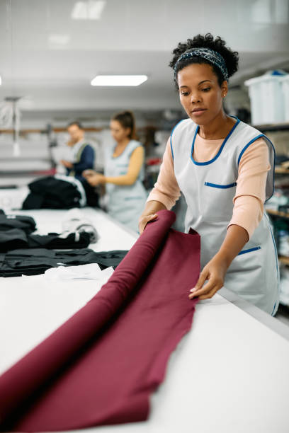 Black female textile worker using new fabric roll while working at tailor's workshop. Young African American seamstress working with fabric at clothing design workshop woman tailor stock pictures, royalty-free photos & images