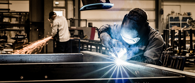 Closeup of heavy industry workers. One of the workers is welding two pieces of metal with arc welding machine. The guy in focus is grinding what his colleague just welded. He's using electric grind stone machine. Deep blue toned shot.