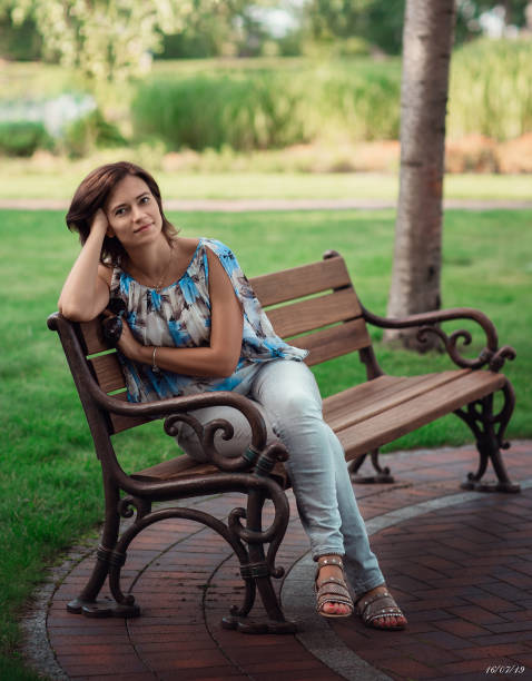 женщина 40 лет сидит на скамейке в парке. лето, деревья, тишина.
женщина в джинсах и блузке. средний возраст - 35 40 years стоковые фото и изображения