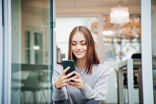 Happy smiling young woman sitting and relaxing on metal stairway in front of modern office cafeteria entrance reading the news and social media messages in her free time on her Smart Phone.