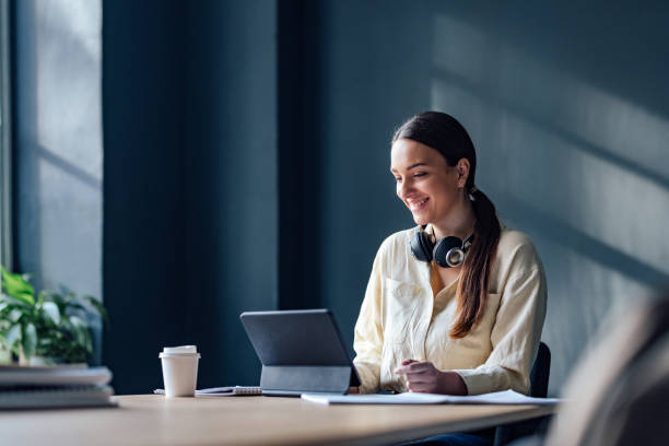 Happy Female Student Studying Online On A Digital Tablet Smiling woman sitting at desk and reading online lesson on her tablet in the library. e learning stock pictures, royalty-free photos & images