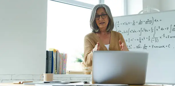 Photo of Mature woman teaching mathematics while standing near the whiteboard and looking at laptop