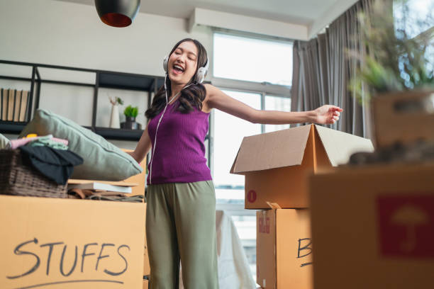 ้幸福 アジアの女性 大人の女性 陽気なダンスの動きと箱から物を開梱 リラックス カジュアルにヘッドフォンを身に着けながら 新しい家、家の家のアパート ブームのアイデアの概念 - 家 掃除 ダンス ストックフォトと画像
