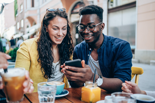 Shot of multiracial group of young people sitting in a sidewalk cafe using a phone, talking, drinking coffee, smiling and having fun.