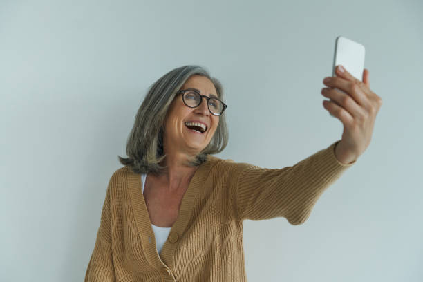 istock Happy senior woman making selfie by her smart phone while standing against white wall 1409886514