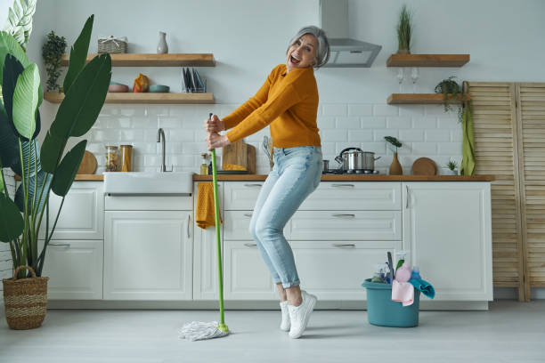 playful senior woman dancing with a mop while standing at the domestic kitchen - spring cleaning women cleaning dancing imagens e fotografias de stock