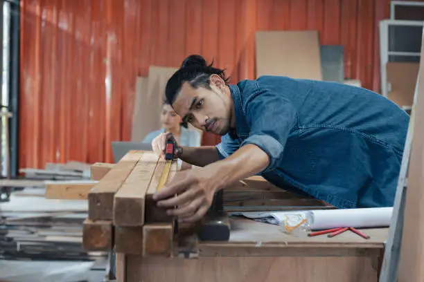 Photo of Young Asian man Carpenter uses a tape measure to measure wood on the workbench in woodcraft carpentry workshop.