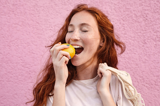 Portrait view of the red haired woman biting tasty orange at the street over pink urban wall. Healthy food and summer concept