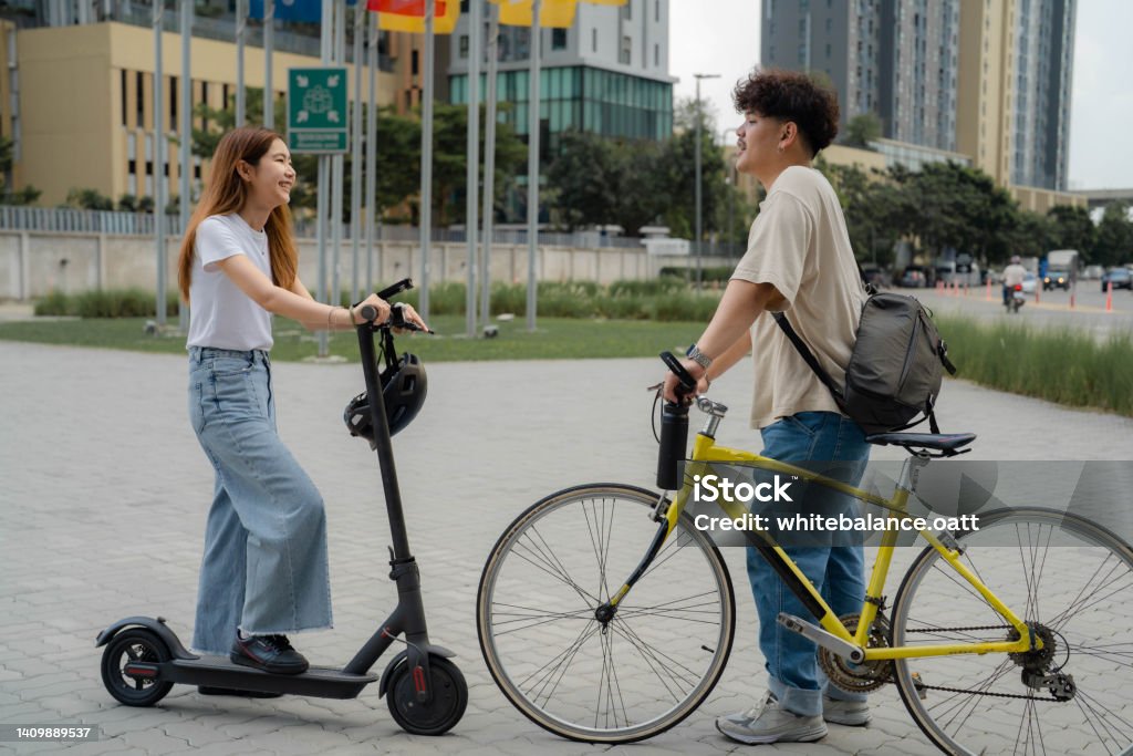 Two young freelancer commuting with environmentally friendly vehicles. Attractive Asian woman riding e-scooter greeting and talking with her coworker riding bicycle on the way go to their office in the morning. Bicycle Stock Photo