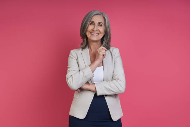 Confident senior woman in formalwear smiling while standing against pink background stock photo