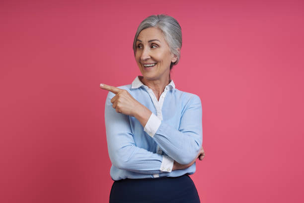istock Confident senior woman pointing away and smiling while standing against pink background 1409890217