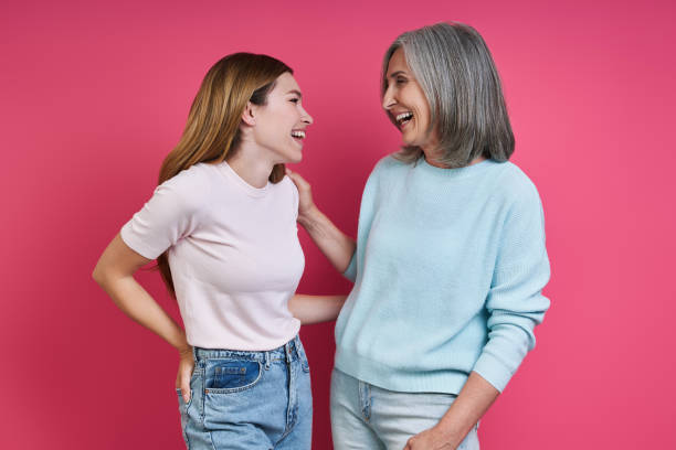 Happy mother and adult daughter looking at each other against pink background stock photo