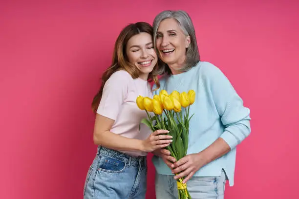 Photo of Happy senior mother and adult daughter holding a bunch of tulips against pink background