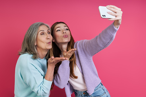Happy senior mother and adult daughter making selfie against pink background