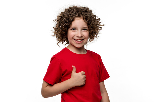Positive Ñurly short-haired girl 10-12 years old wears red Basic t-shirt. Friendly preteen schoolgirl showing like, approval sign, thumb up standing on white background studio.