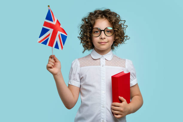 Girl holds book and UK flag Child Curly-haired schoolgirl 10-12s in white blouse and round glasses looking at camera. Girl holds red textbook and shows UK British flag on light blue studio background. Study abroad concept Study Abroad Countries stock pictures, royalty-free photos & images