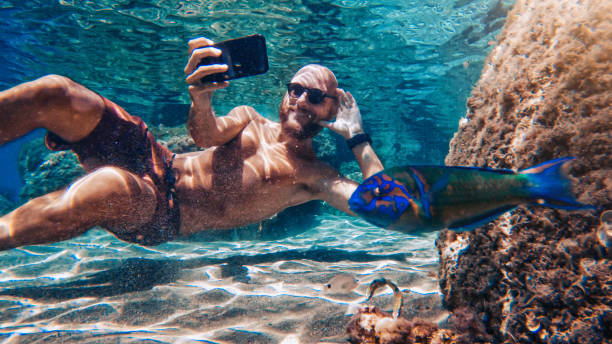 Selfie with mobile phone underwater at sea: fish photobombing Funny man selfie with mobile phone underwater at sea: internet addiction. photo bomb stock pictures, royalty-free photos & images