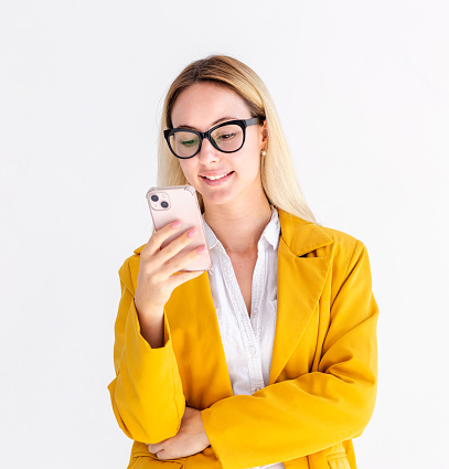 Young businesswoman in a jacket holding a mobile phone, typing a message, online chatting. Female wearing eyeglasses looks at smartphone screen and smiling standing on a white background