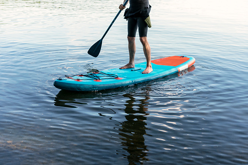 Man faceless in thermo suit paddling stand up paddleboard blue SUP on the water lake with paddle male legs active lifestyle summer activities copy space