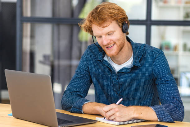 istock Happy male call center worker with headset consults a client. Redhead man freelancer or student using laptop for remote work from home. Learning, education remotely online, studying 1409893811