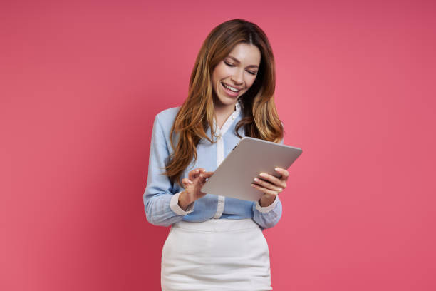 istock Beautiful young woman using digital tablet and smiling while standing against colored background 1409895476