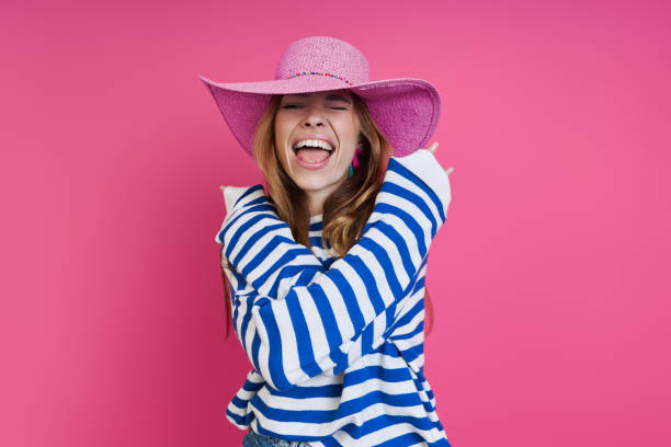 Excited young woman in pink hat hugging self while standing against colored background