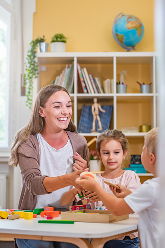 Kindergarten teacher playing together with children at colorful preschool classroom. Mother playing with children.