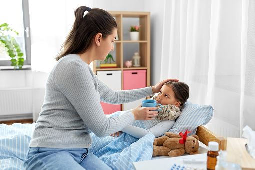 family, health and people concept - mother giving hot tea sick daughter lying in bed at home