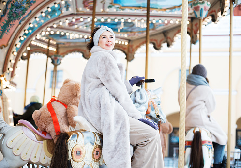Charming woman in a faux fur coat and mittens rides a carousel and has fun