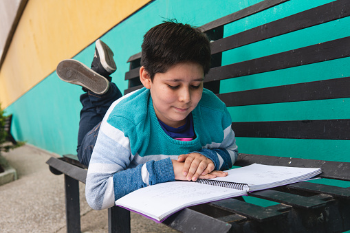 Caucasian boy with his black hair wearing a sweater on a park bench next to a colorful wall with an open book studying very intently