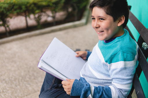 A black-haired Caucasian boy wearing a striped sweater is holding an open book and smiling happily on a park bench. studious child