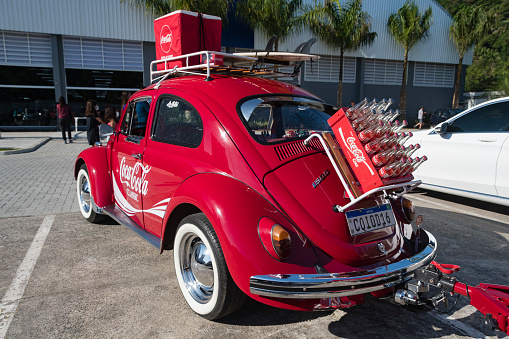 Praia Grande, São Paulo - Brazil - July 25, 2021: Old red Volkswagen Beetle customized with the Coca Cola brand. Luggage compartments with surfboard, chrome hubcaps and white tire strip.