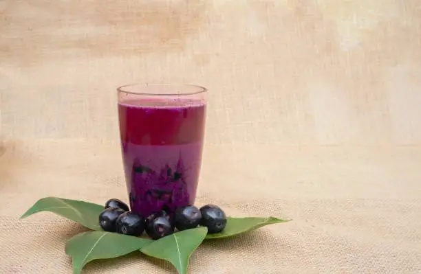 Closeup of Jamun or Syzygium Cumini Juice in a Glass with Fruit and Leaves Isolated on Burlap Fabric with Copy Space, Also Known as Java Plum, Malabar Plum, Black Plum or Jambolan in Horizontal Orientation.