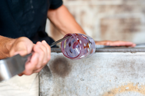 Cloe up of a glass blower shaping melting glass into a glass by turning it and using a plier. Focus on the glass rim even if the shot is motion blurred.