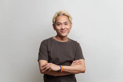 Studio portrait photo of young Asian man wearing rainbow wristband with arms crossing on white background. Young man emotion face expression portrait concept.
