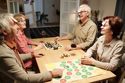 Four cheerful aged friends with notepapers on their foreheads sitting around table with chessboard and paperboard and playing name game