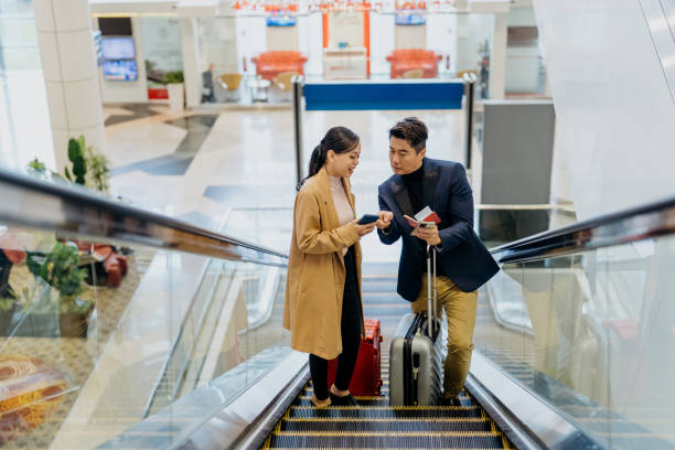Two Asian business travellers riding on escalator in the airport Image of an Asian Chinese businessman and Malay businesswoman riding one escalator to airport departure area for business trip klia airport stock pictures, royalty-free photos & images