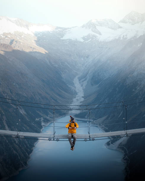 hombre con chaqueta amarilla sentado en un puente colgante sobre un lago en olpererhutte, austria - sports danger fotografías e imágenes de stock