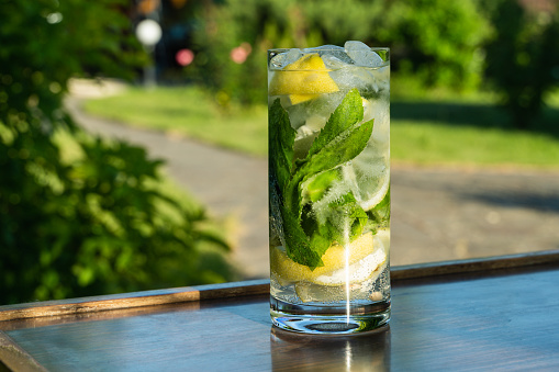 Cold iced mineral water with mint leaves and lemon pieces in tall glass on wooden table on outdoor background.