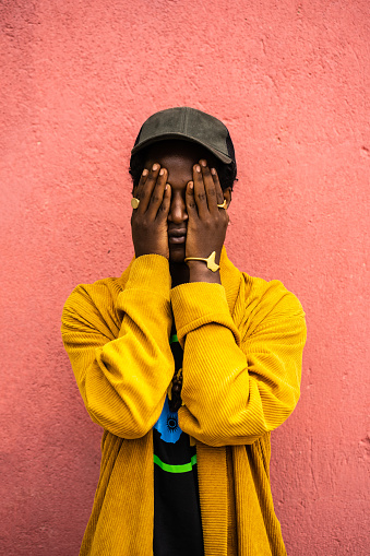 African man standing against orange wall with both his hands touching and covering his face