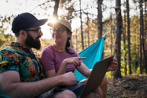 Couple balancing between vacation and work sitting on a hammock using laptop on camping vacation in nature.