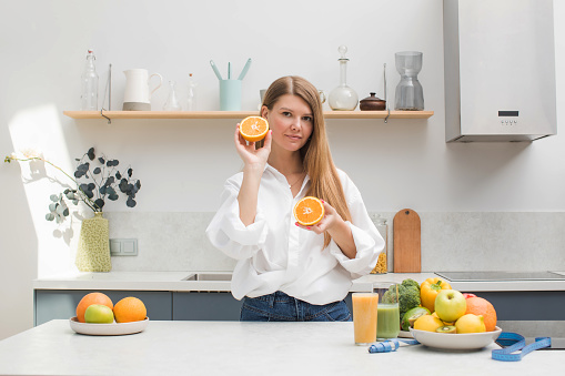 A young blond woman holds orange halves in her hands in the kitchen next to a table with fruits. The concept of diet, healthy eating.