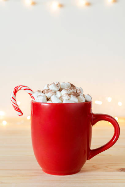 istock Hot chocolate or cacao in red mug with small marshmallows and candy cane on table with holiday lights on wall. 1409952870