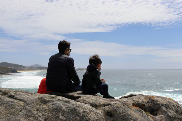 istock Father and young son sitting on a rock looking out to sea. 1409962017