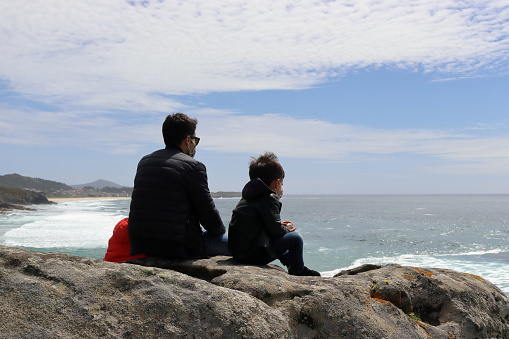 Father and young son with jackets on sitting on a rock looking out to sea.