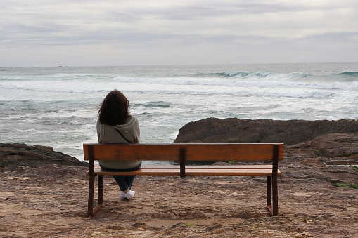 WOMAN SEEN FROM BEHIND, SITTING ON THE LEFT, ON A WOODEN BENCH WHILE CONTEMPLATING THE SEA.