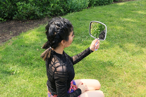 An East Indian model checking herself in a handheld mirror. She is wearing a black long sleeved see through shirt and a multi colored mini skirt.
